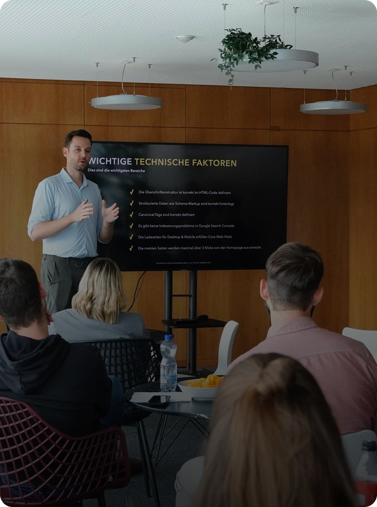 Ein Mann steht vor einem großen Bildschirm und hält vor einem sitzenden Publikum in einem modernen Konferenzraum einen Vortrag über technische Faktoren.